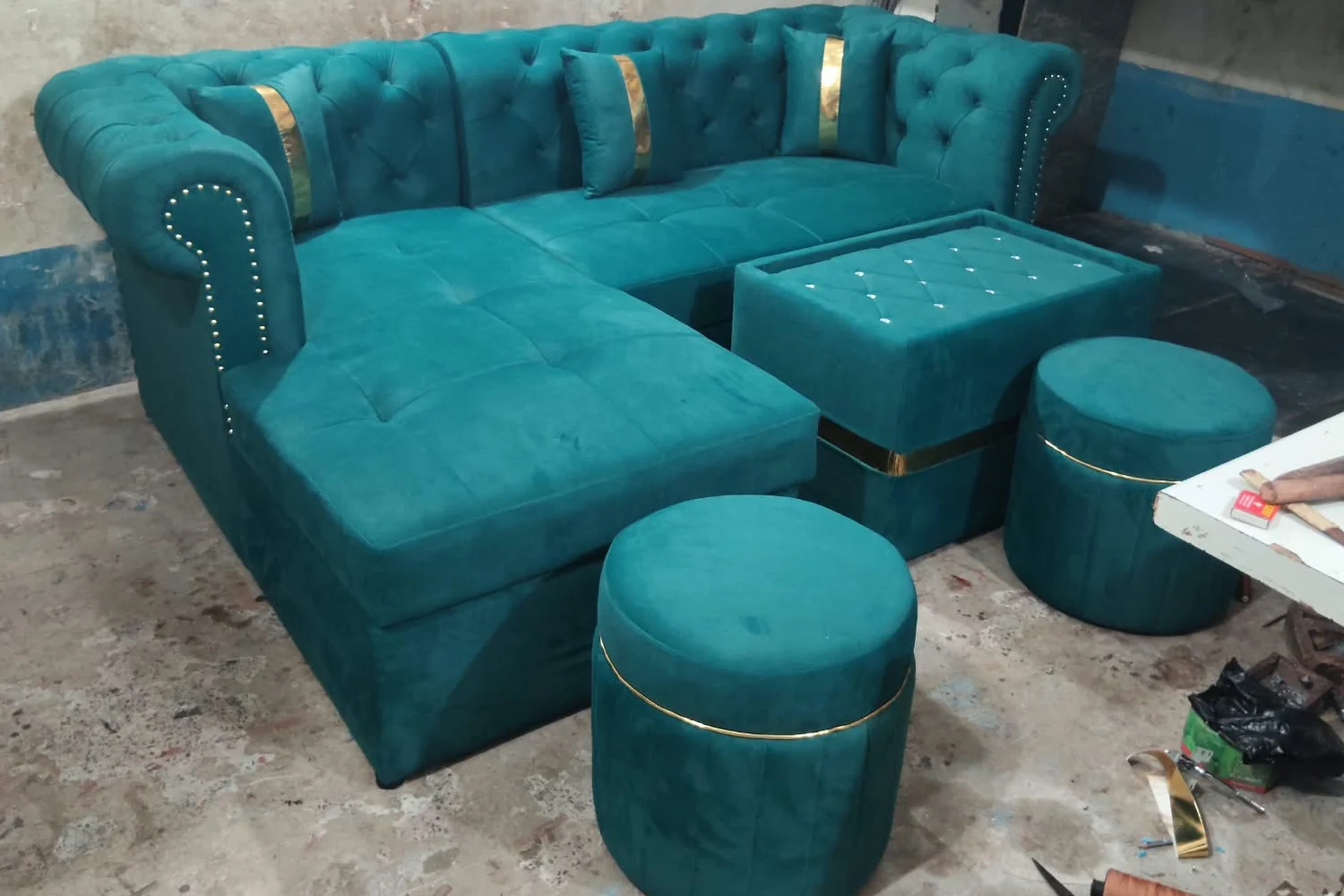 An emerald buttoned chesterfield corner set with gold trim, matching ottomans and a centre table, finished on the Afia workshop floor beside the workbench.
